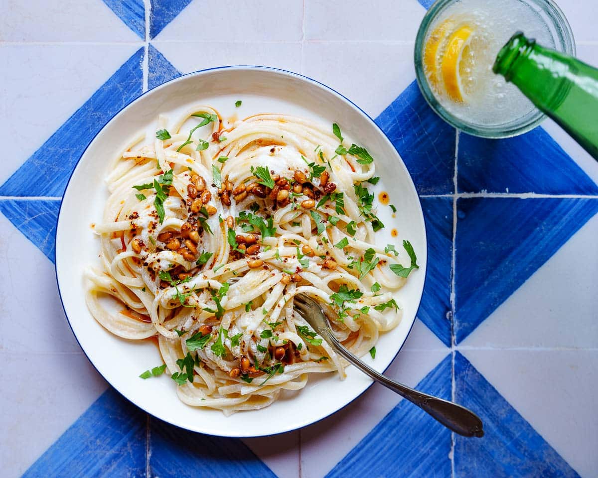 Linguine i en hvit saus med pinjekjerner og bladpersille på toppen, på en hvit asjett med blå kanter på blå/hvitrutet bakgrunn. I kanten av bildet helles farris fra en grønn flaske i et glass med sitronskiver i. Bildet er tatt ovenfra.