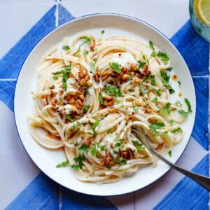 Linguine i en hvit saus med pinjekjerner og bladpersille på toppen, på en hvit asjett med blå kanter på blå/hvitrutet bakgrunn. I kanten av bildet skimtes et glass med farris og sitronskiver. Bildet er tatt ovenfra.