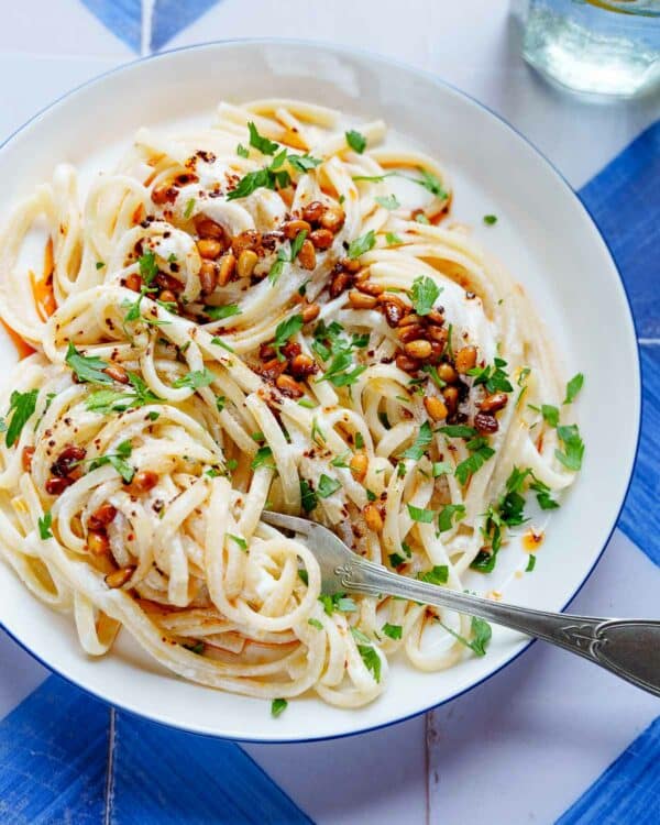 Linguine i en hvit saus med pinjekjerner og bladpersille på toppen, på en hvit asjett med blå kanter på blå/hvitrutet bakgrunn. I kanten av bildet skimtes et glass med farris og sitronskiver. Bildet er tatt fra øyehøyde