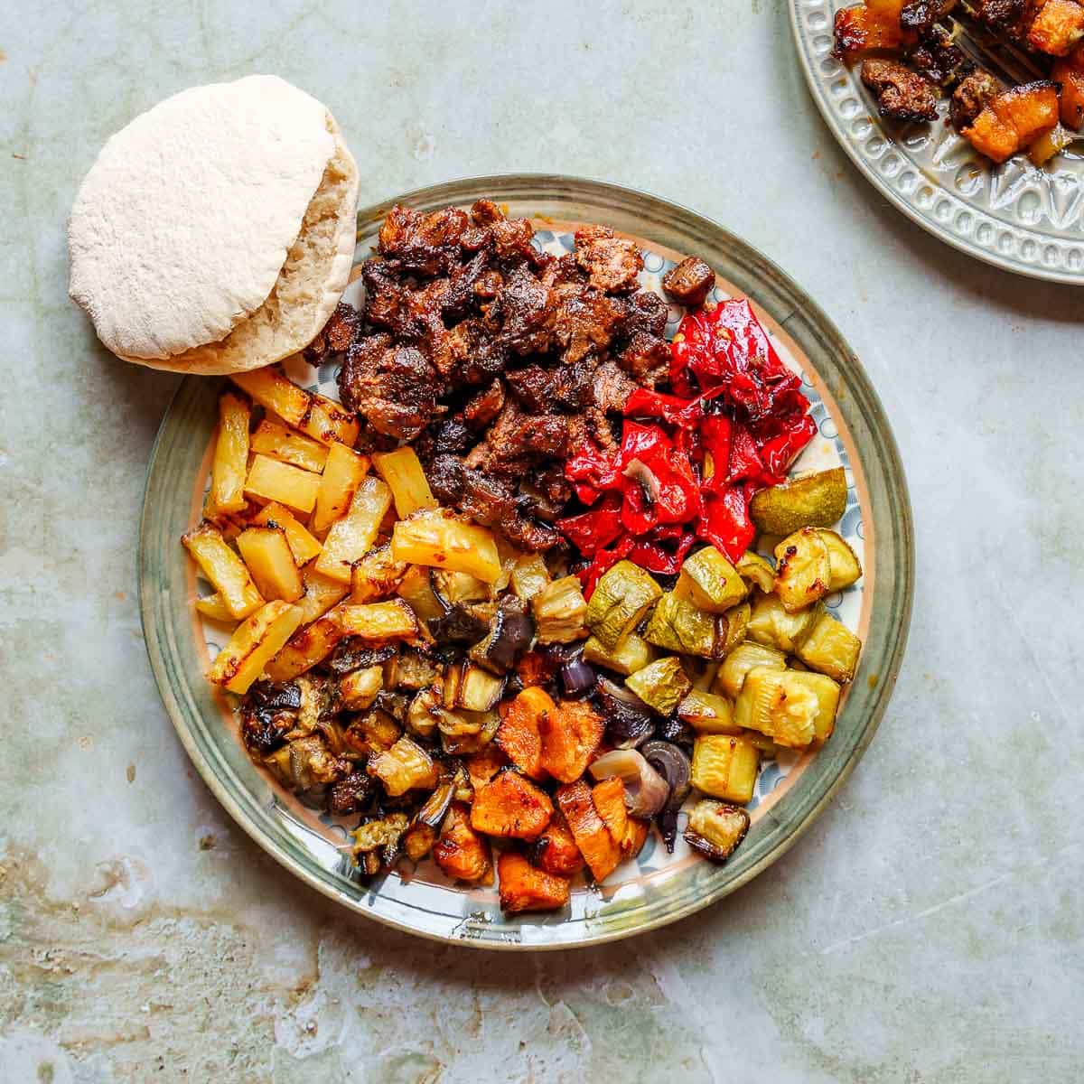 Stor tallerken med stekt lammekjøtt, potet, aubergine, søtpotet. løk, squash og paprika side ved side. Et pitabrød hviler på kanten av fatet, som står på et olivengrønt marmorbord og ses ovenfra.